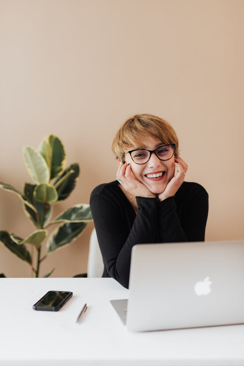 Social Media 1 Cheerful woman in eyeglasses and black sweater smiling while working at a desk with a laptop indoors.