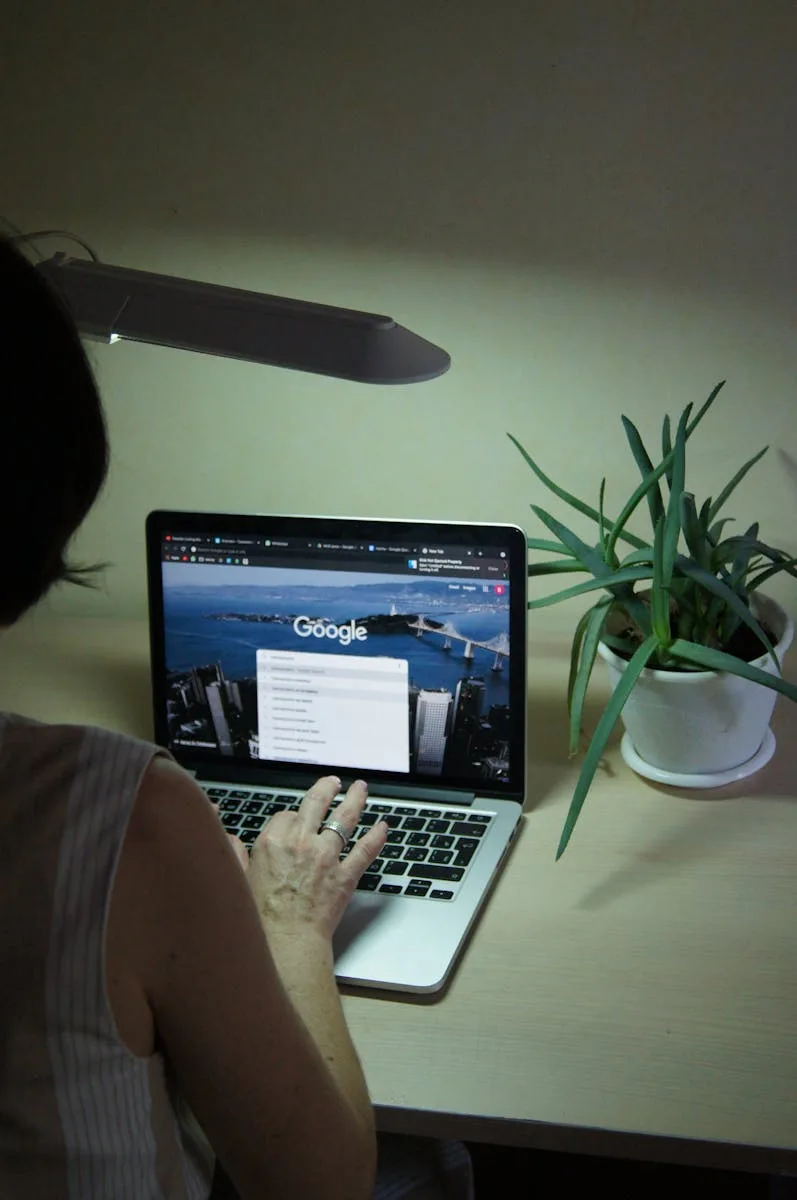 An adult browsing online with a laptop on a desk with a plant nearby, under soft lighting.