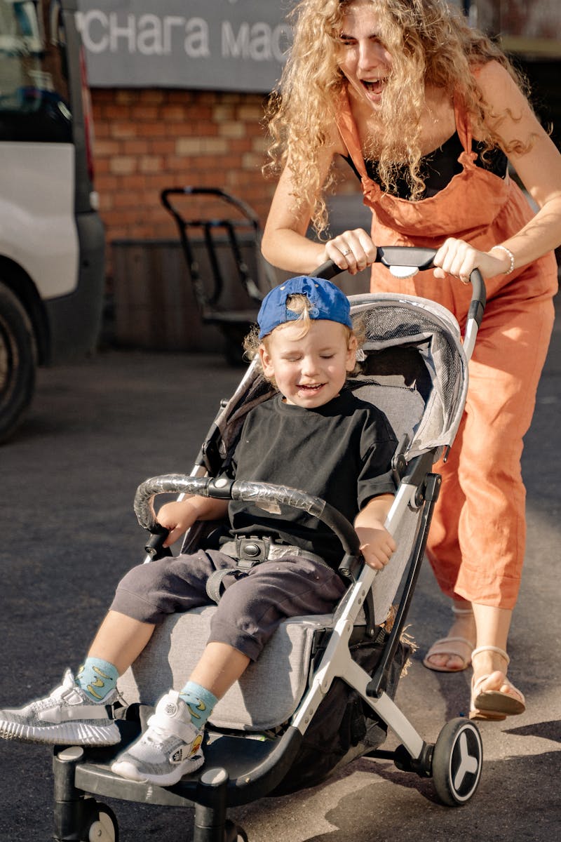 A mother joyfully pushes her smiling son in a stroller on a sunny day.