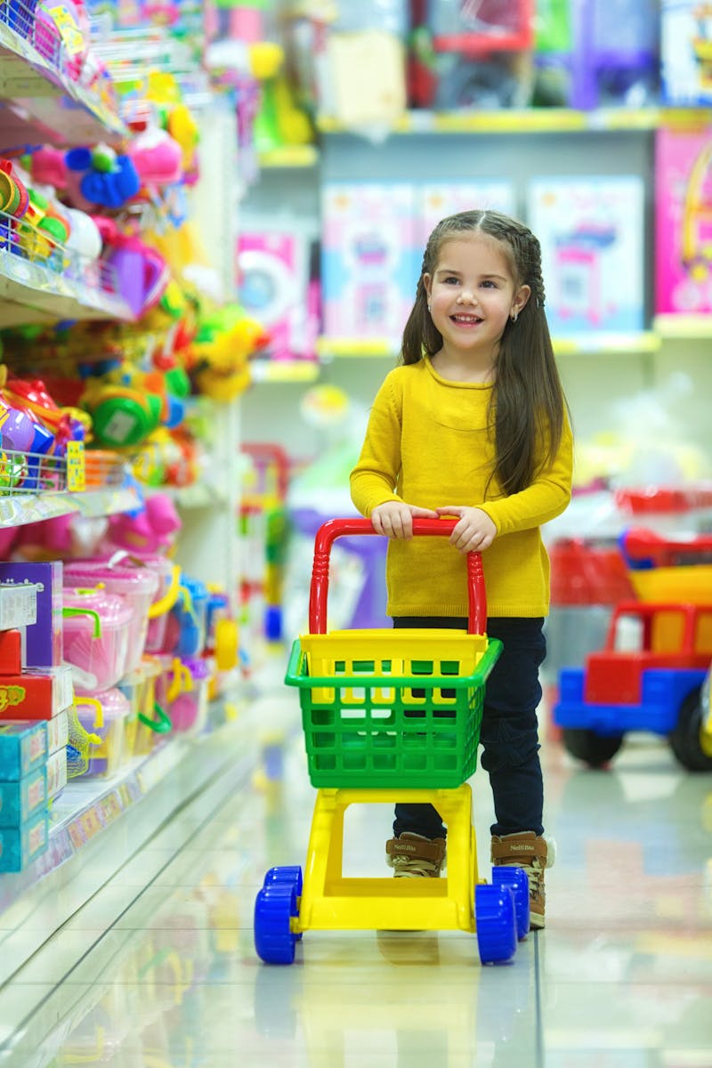 Adorable young girl happily pushing a toy shopping cart in a colorful toy store aisle.
