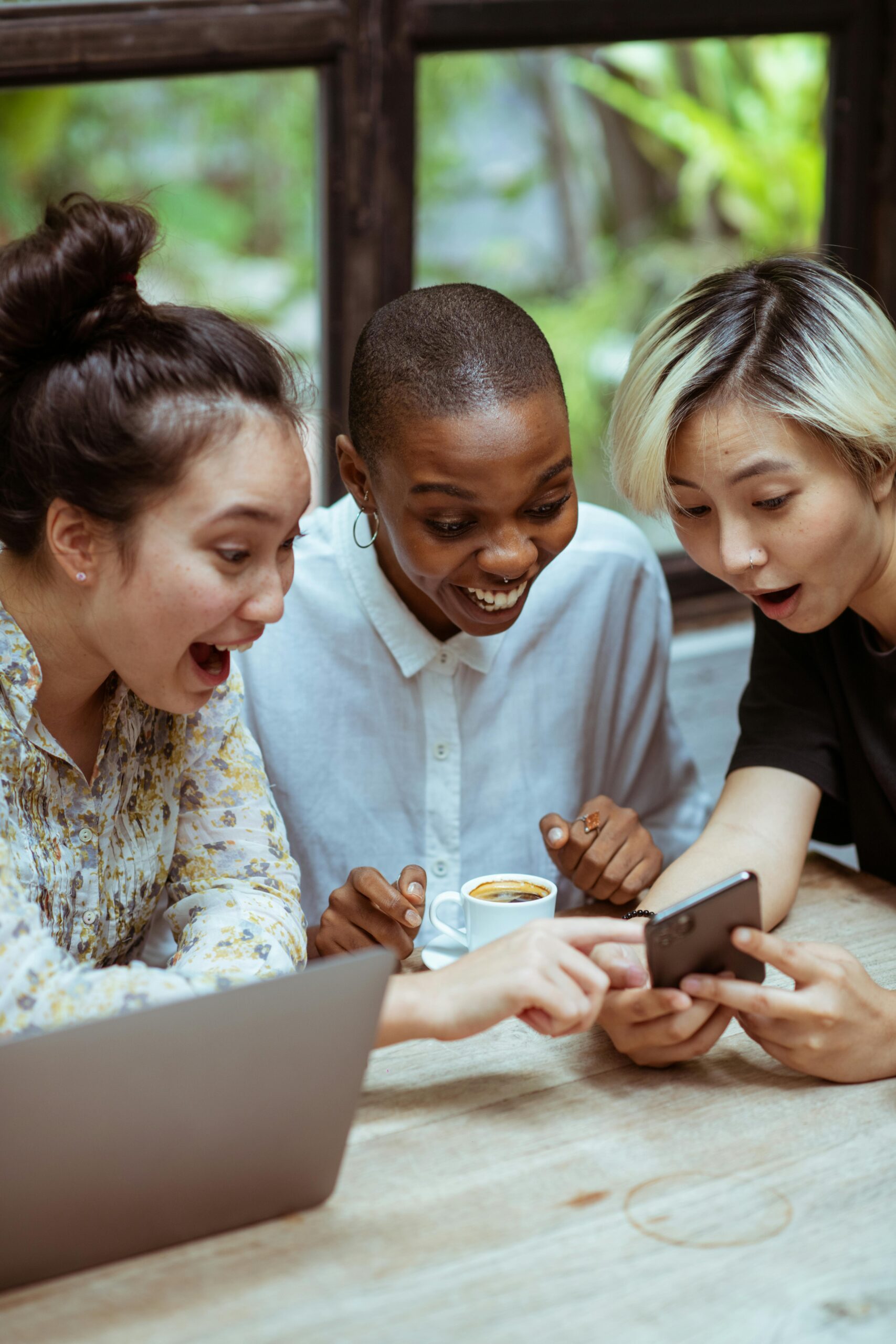Social Media Marketing for Children's Brands 2 Amazed surprized multiethnic female friends in casual clothes with cup of coffee browsing smartphone while sitting at wooden table with laptop against window