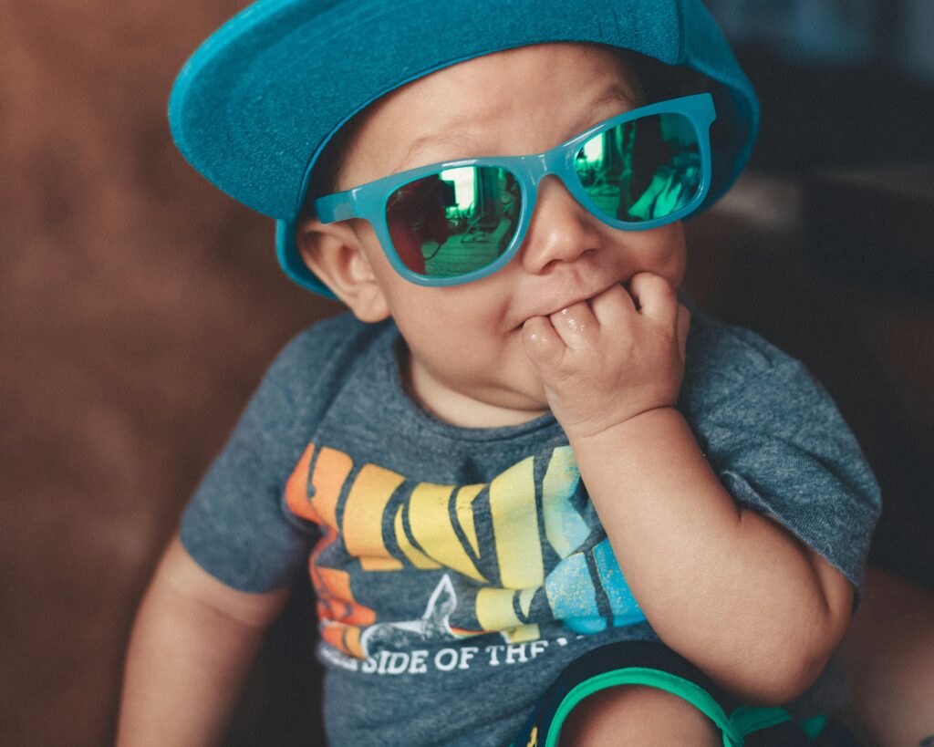 Adorable baby wearing sunglasses and a cap, sitting playfully indoors.