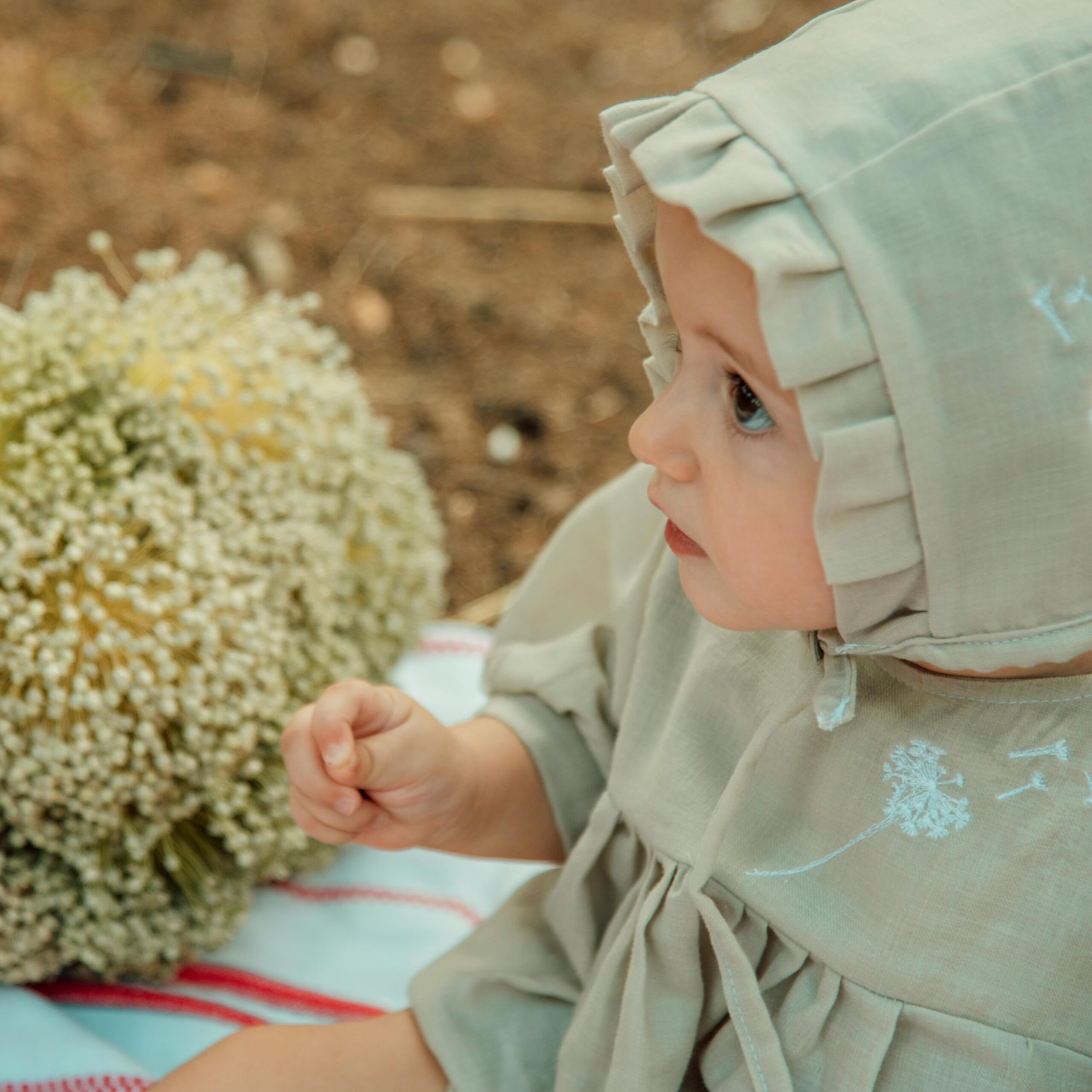 Charming baby in hooded outfit sitting beside a bouquet in nature setting.