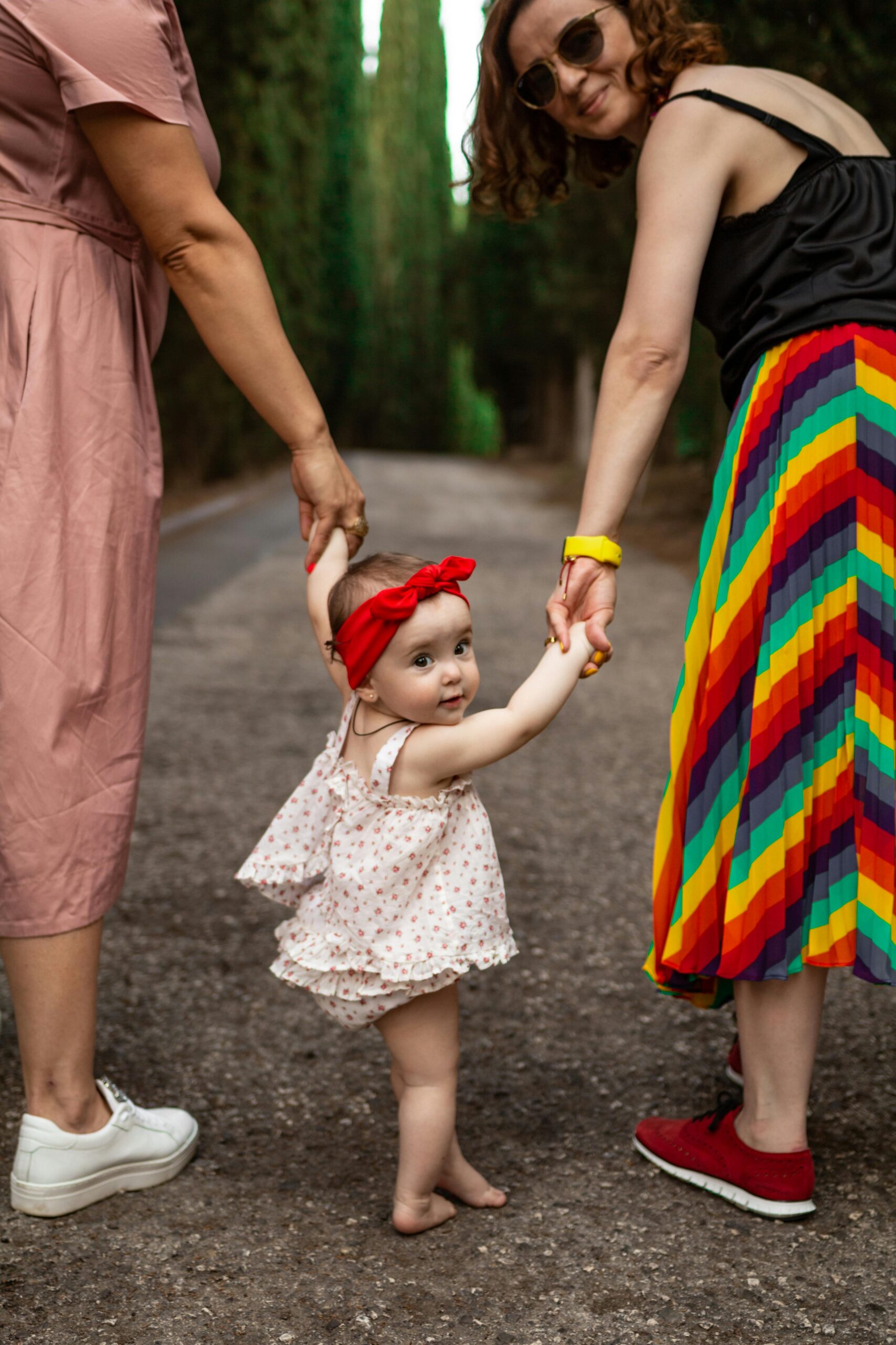 A cute baby wearing a bow walks with her mother in a colorful skirt during summer.