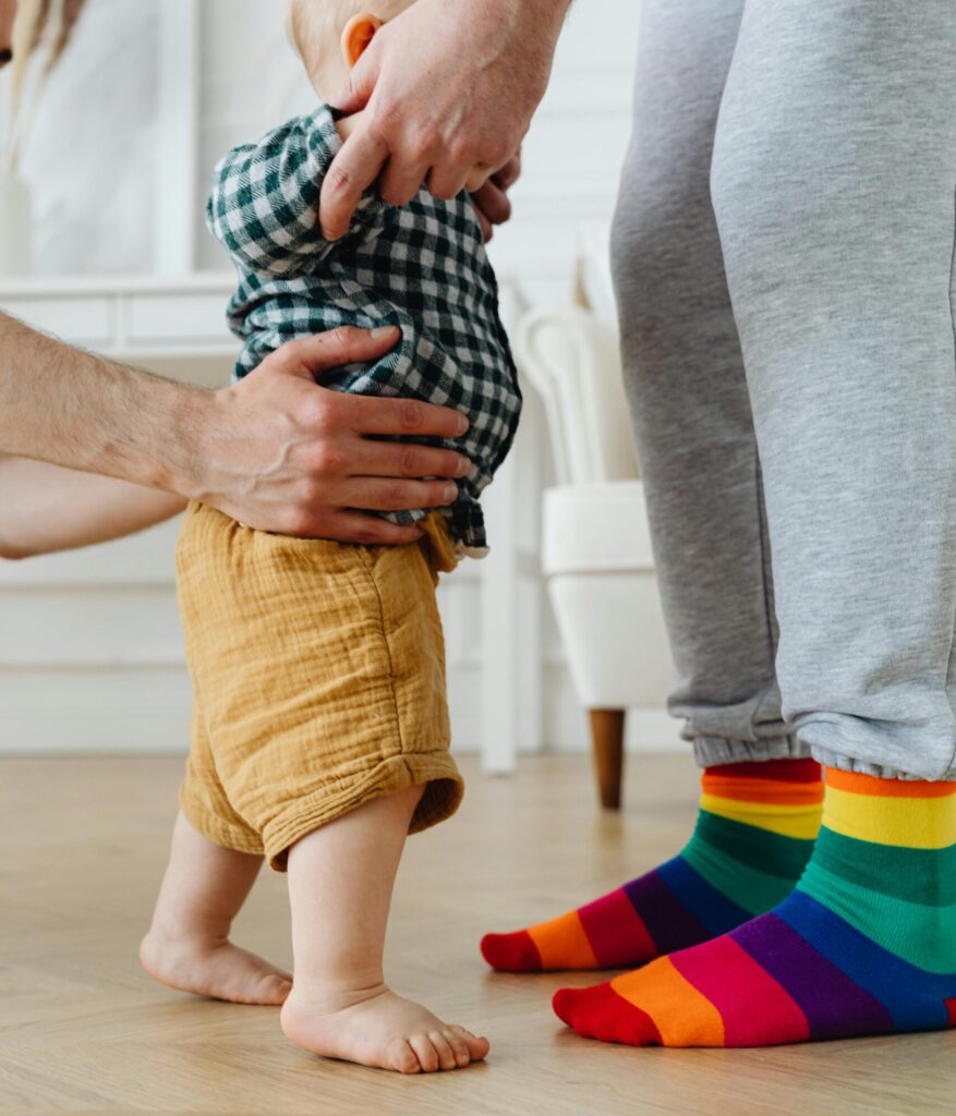 An adorable baby is taking its first steps indoors, supported by a parent. Colorful socks add a playful touch.