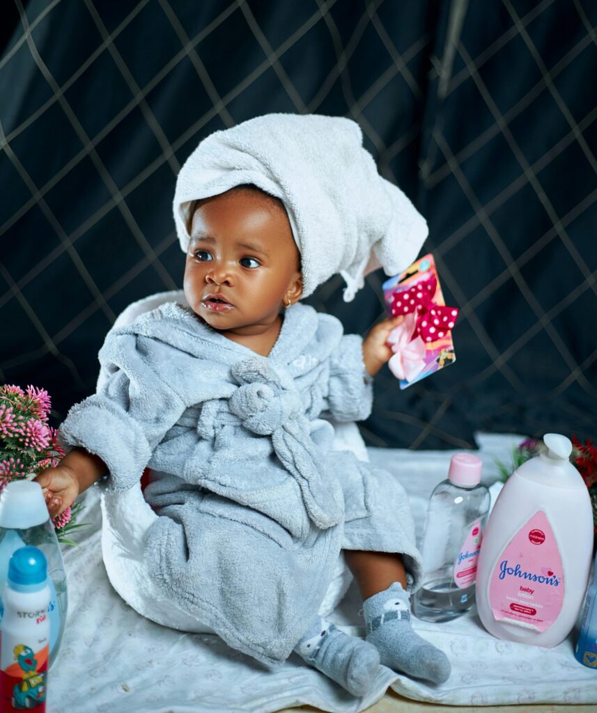 Cute baby in a bathrobe surrounded by skincare products, indoors.