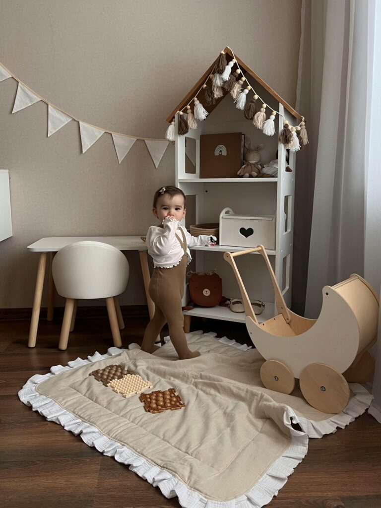 A baby stands in a stylish playroom with a wooden stroller and toys, evoking warmth.