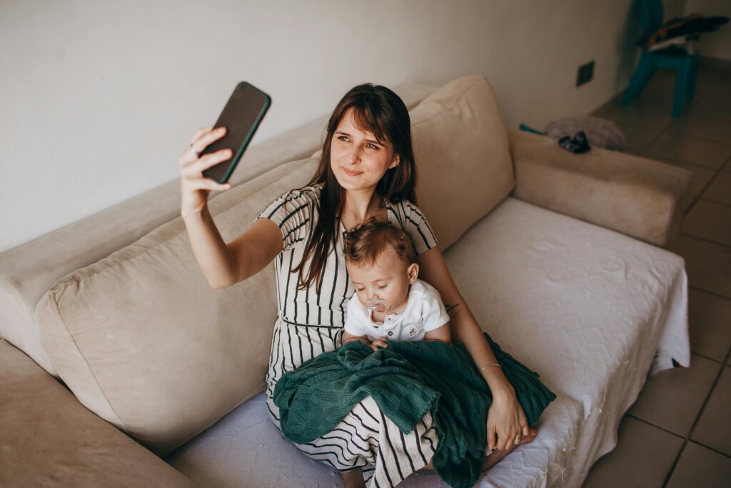 Smiling mother takes a selfie with her baby sitting on a cozy sofa indoors.
