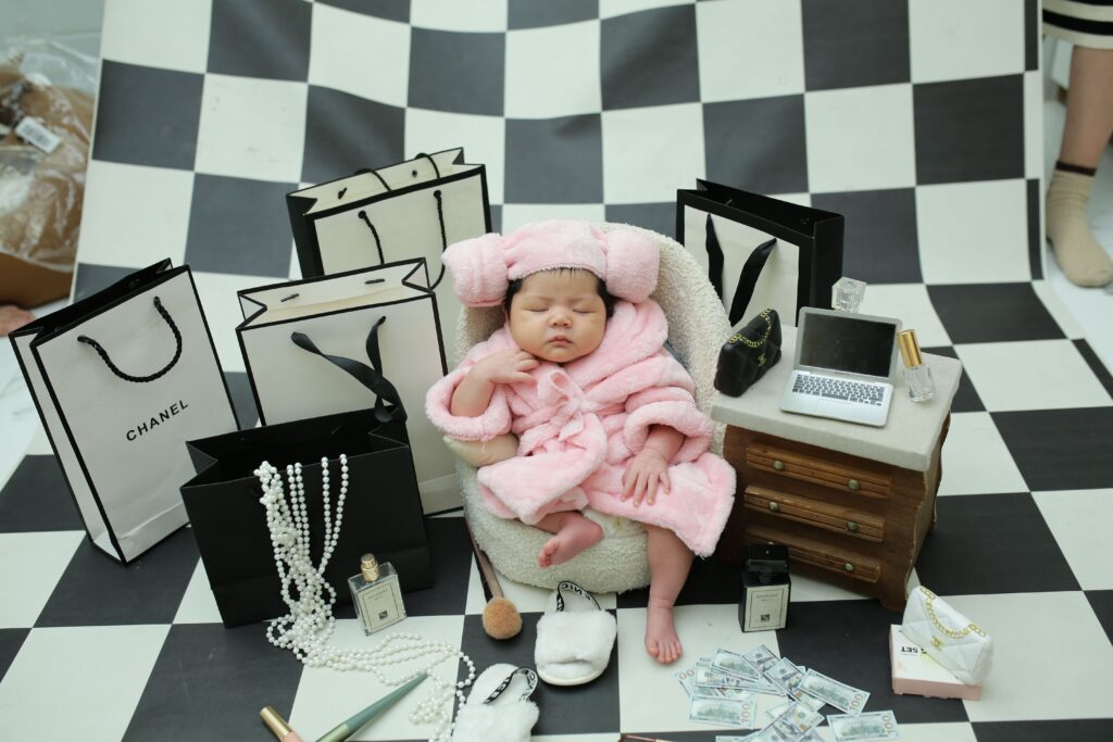Charming baby dressed in pink surrounded by luxury shopping bags and accessories.