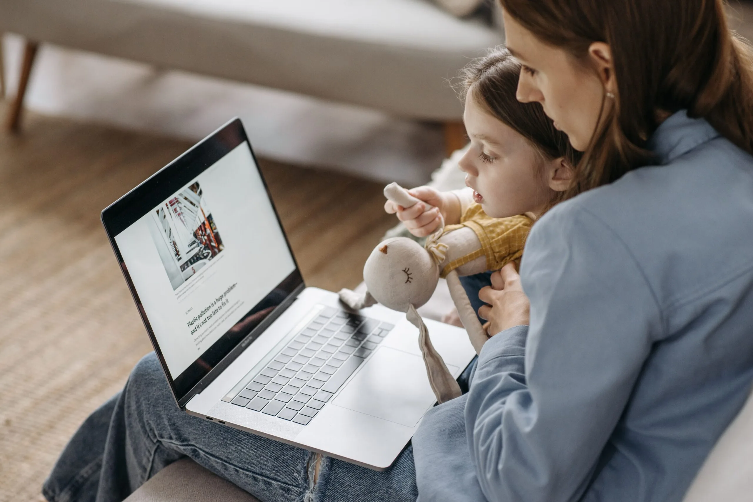 A mother works from home on a laptop with her daughter sitting on her lap, holding a stuffed toy.