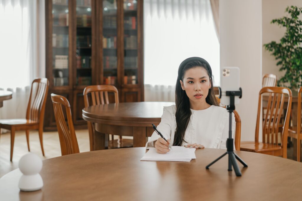 Young Asian woman vlogging in a cozy home office setup using a smartphone and tripod.
