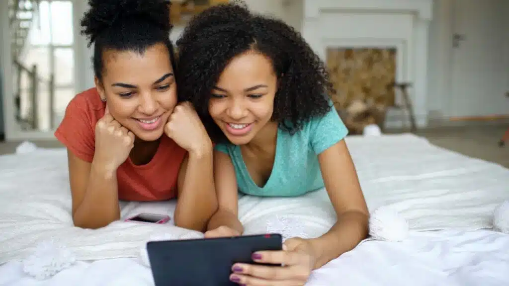 Generación Alfa y marcas infantiles: cómo adaptar marketing, contenido y confianza 12 Two young women lying on bed, smiling and watching a tablet indoors.