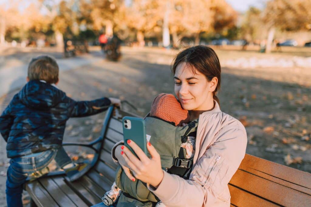 Woman enjoying a sunny day in the park, holding her baby and using a smartphone.