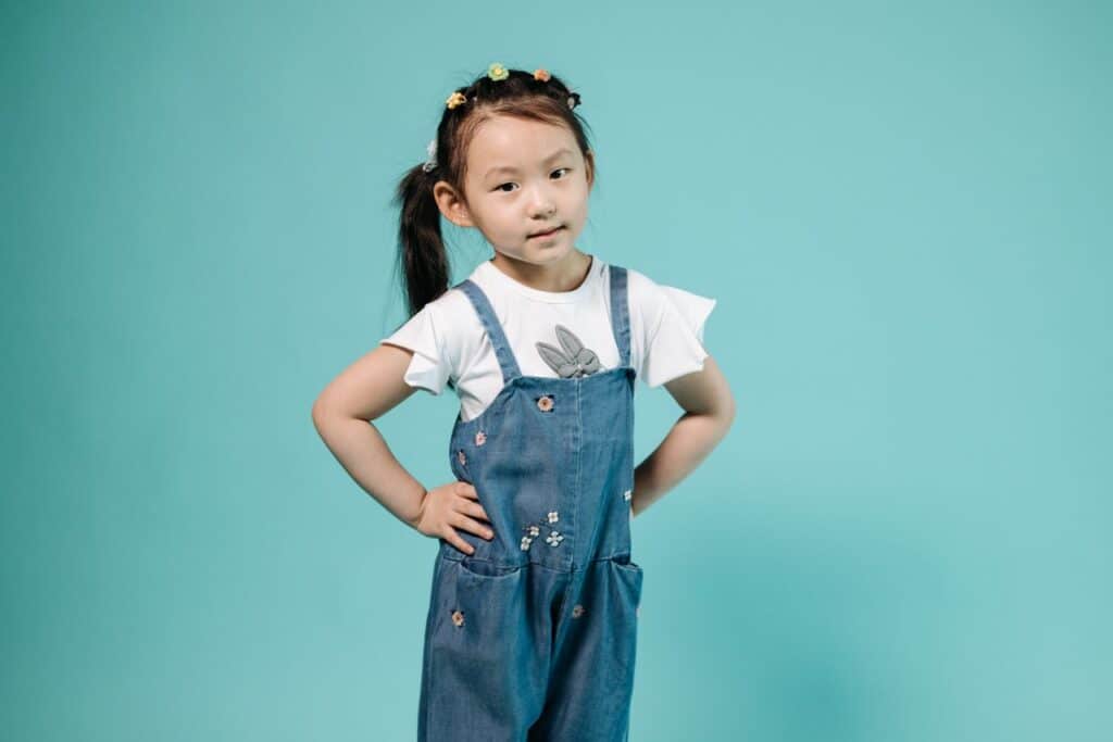 Adorable young girl with hands on her hips posing in a denim overall on a blue background.
