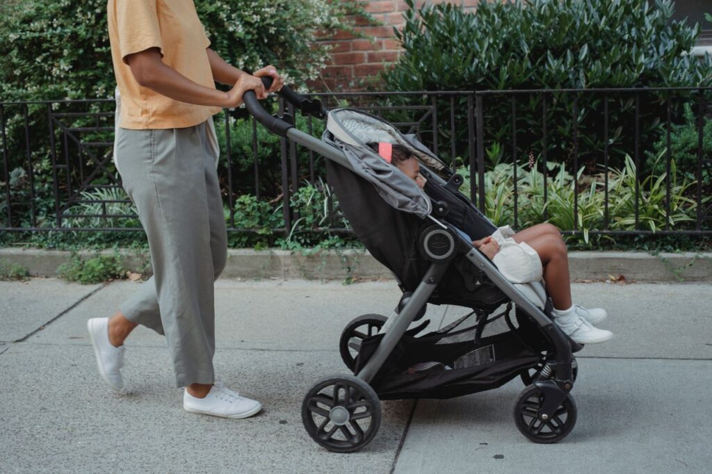 Crop anonymous ethnic female in casual clothes strolling on paved street of city with little girl in carriage
