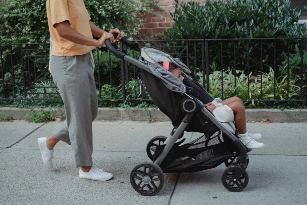 Crop anonymous ethnic female in casual clothes strolling on paved street of city with little girl in carriage