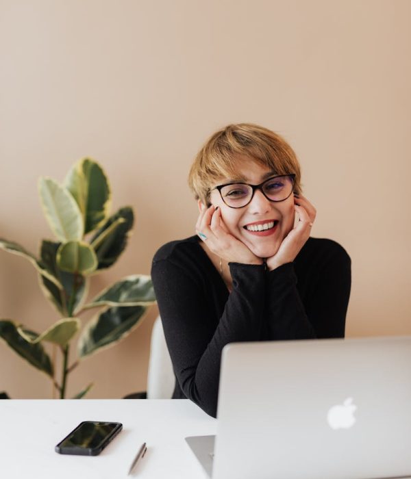 Cheerful woman in eyeglasses and black sweater smiling while working at a desk with a laptop indoors.