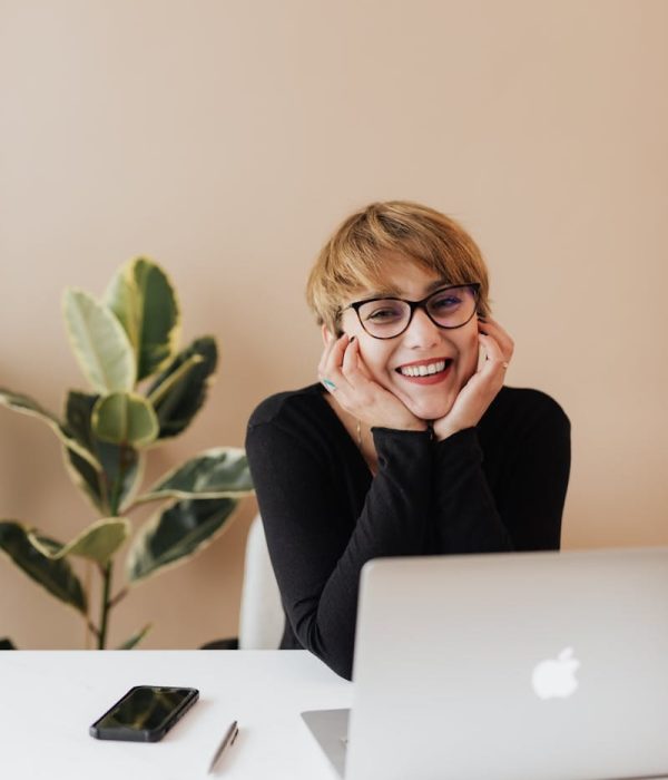 Cheerful woman in eyeglasses and black sweater smiling while working at a desk with a laptop indoors.