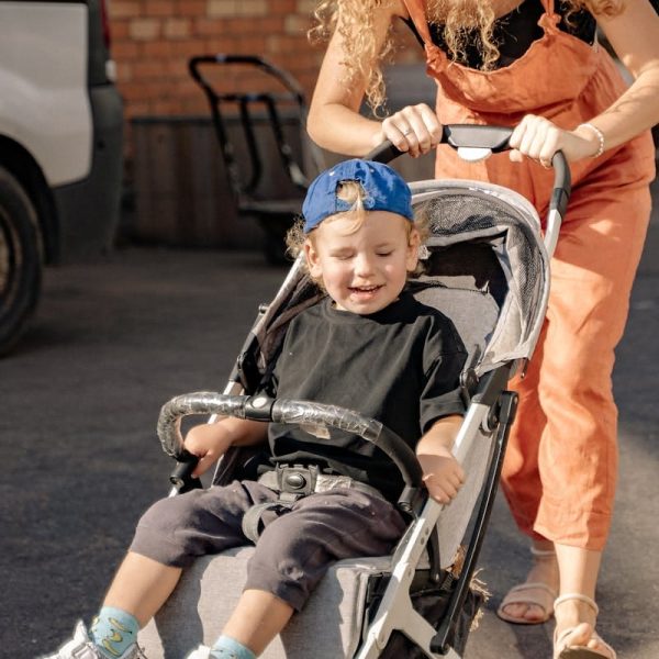 A mother joyfully pushes her smiling son in a stroller on a sunny day.