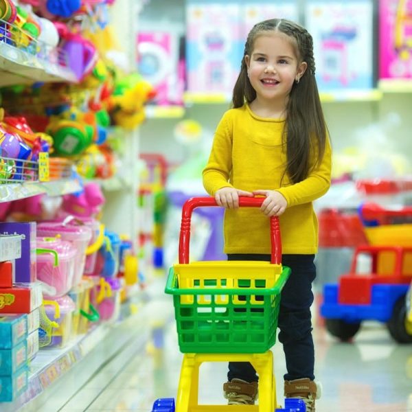 Adorable young girl happily pushing a toy shopping cart in a colorful toy store aisle.