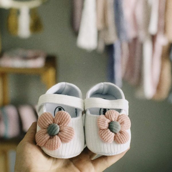 Adorables zapatos de bebé blancos con decoración floral rosa sostenidos en la mano, perfectos para moda de recién nacidos.