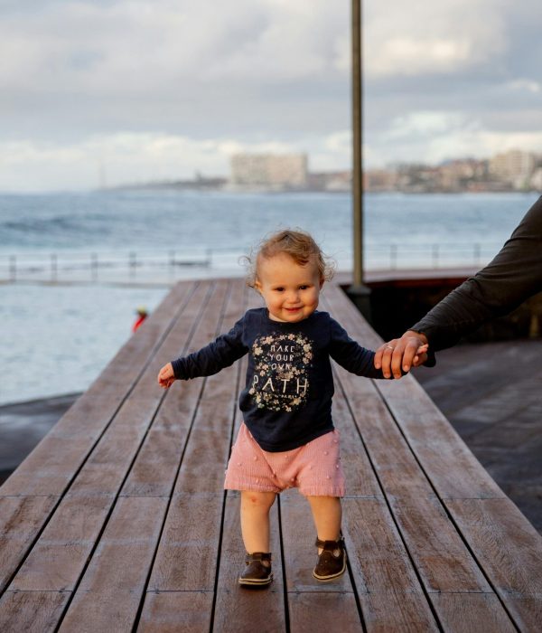 A cute toddler joyfully walks on a wooden dock near the sea while holding an adult's hand. Perfect summer day.
