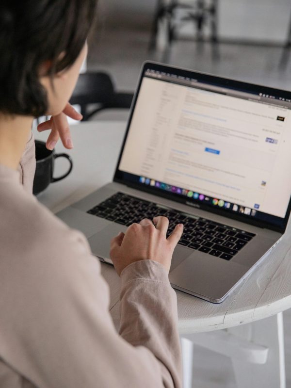 Back view of crop anonymous female remote employee typing on portable computer while watching website on screen and sitting at plastic table with cup of hot drink in apartment