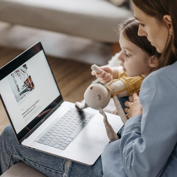 A mother works from home on a laptop with her daughter sitting on her lap, holding a stuffed toy.